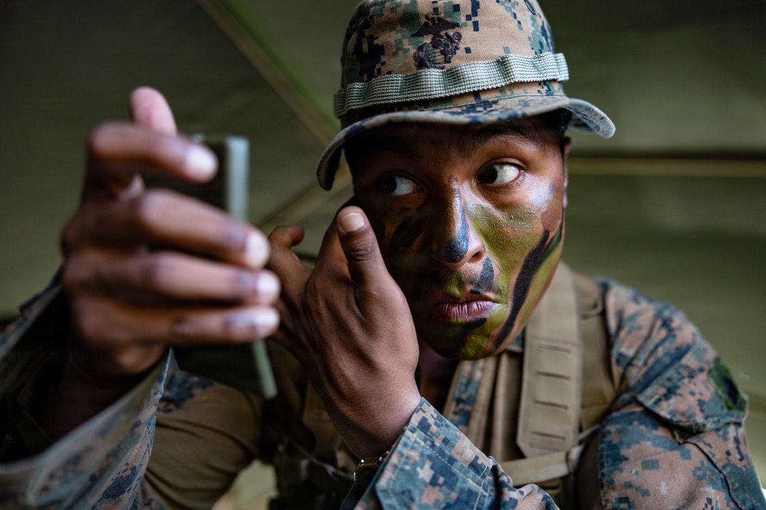 A Marine looks to the left into a compact as they apply camouflage paint to their face.