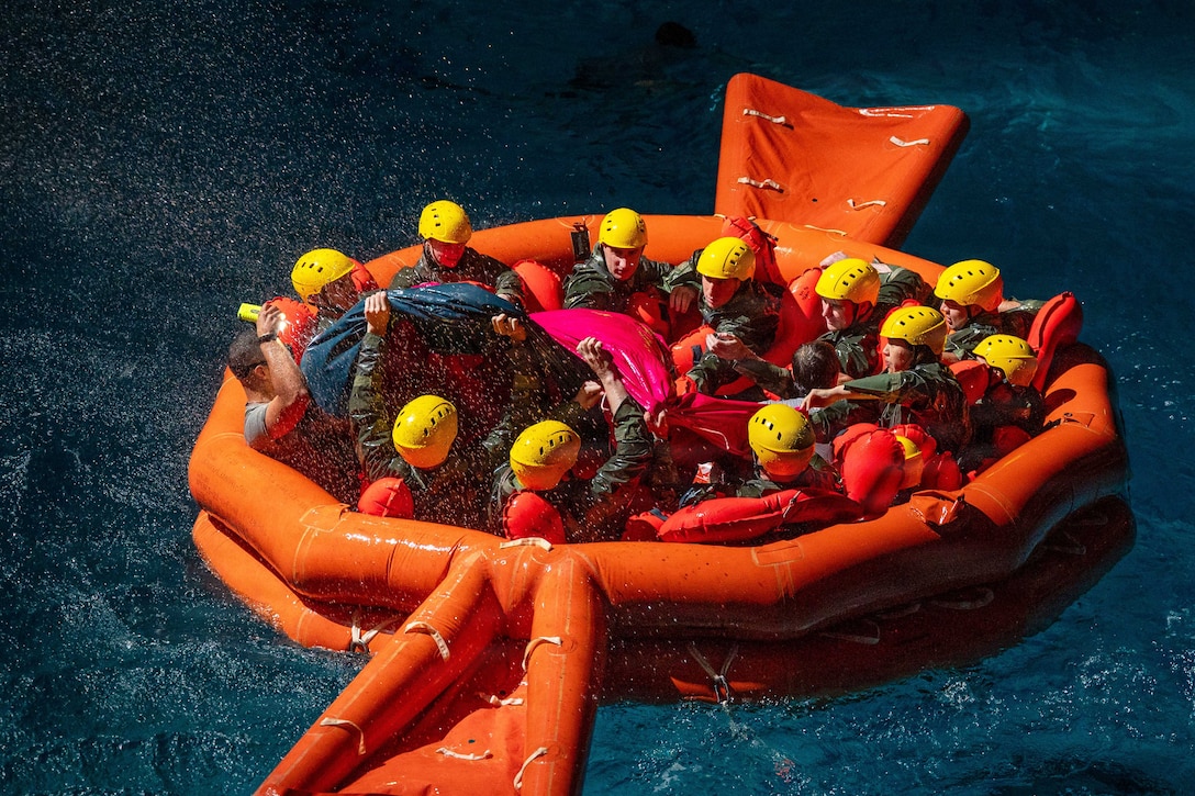 Water splashes as more than a dozen airmen wearing yellow helmets sit in a circular orange raft in a body of water during the day.