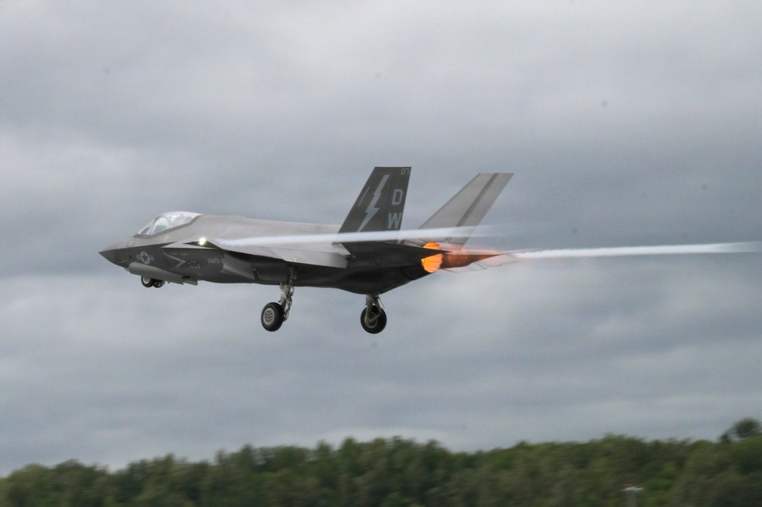 A Marine Corps aircraft flies to the left over trees in a gray sky, leaving behind streaks near an orange engine.