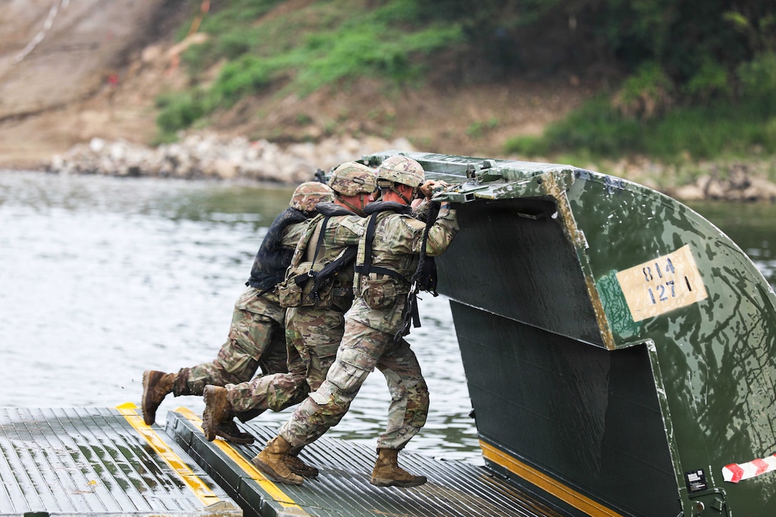 Three soldiers in uniform unfold an improved ribbon bridge in a body of water on a gloomy day.