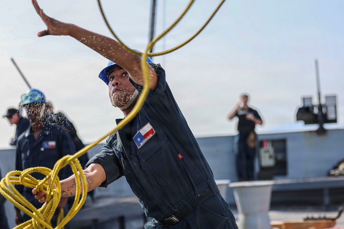 A sailor wearing dark blue coveralls and a blue helmet throws a yellow rope while standing on the deck of a ship on a bright day.