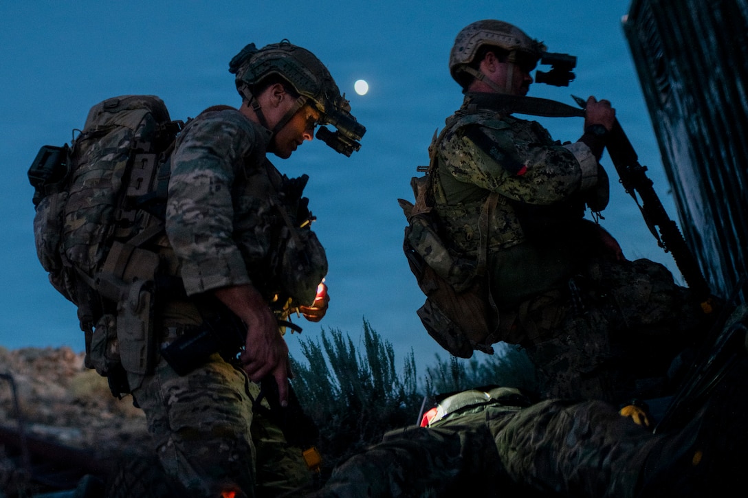 Two sailors wearing camouflage uniforms, helmets with night vision gear, tactical vests and backpacks examine something at night under moonlight.