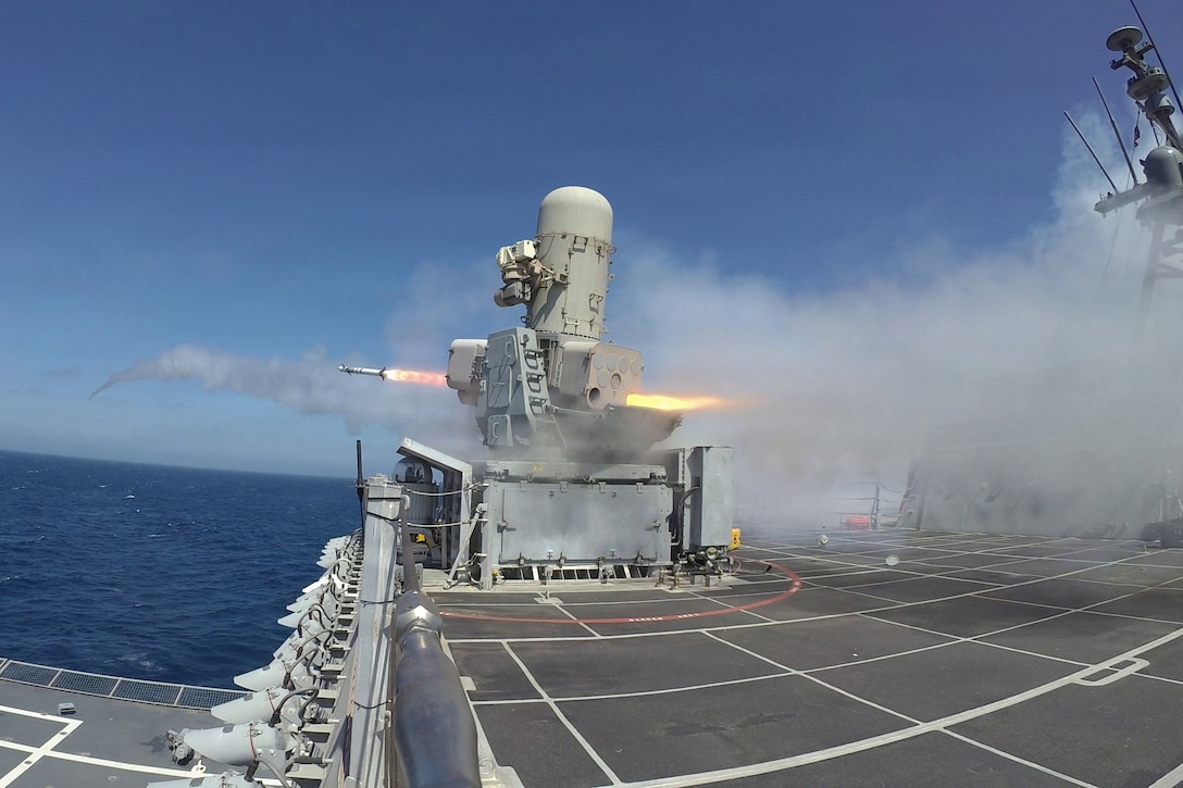 A missile shoots out from a launcher mounted on the desk of a ship on a body of water on a bright day.