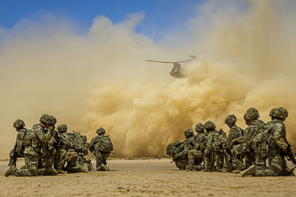 Two groups of about 10 soldiers each kneel as a landing military helicopter kicks up a sand cloud in a desert environment on a bright day.