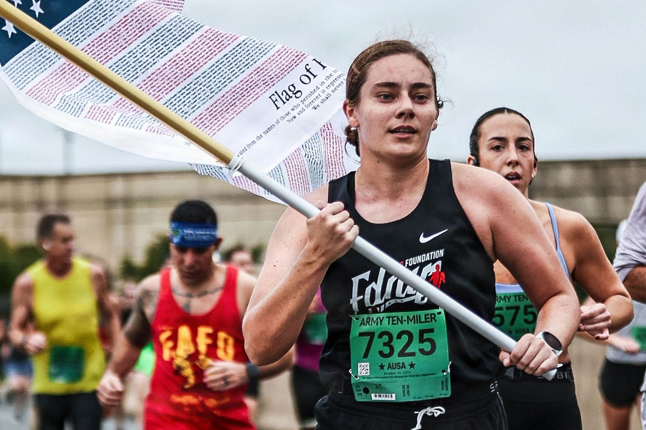 A woman in athletic attire runs a race while carrying a flag. Several other people in similar attire run behind and beside her.