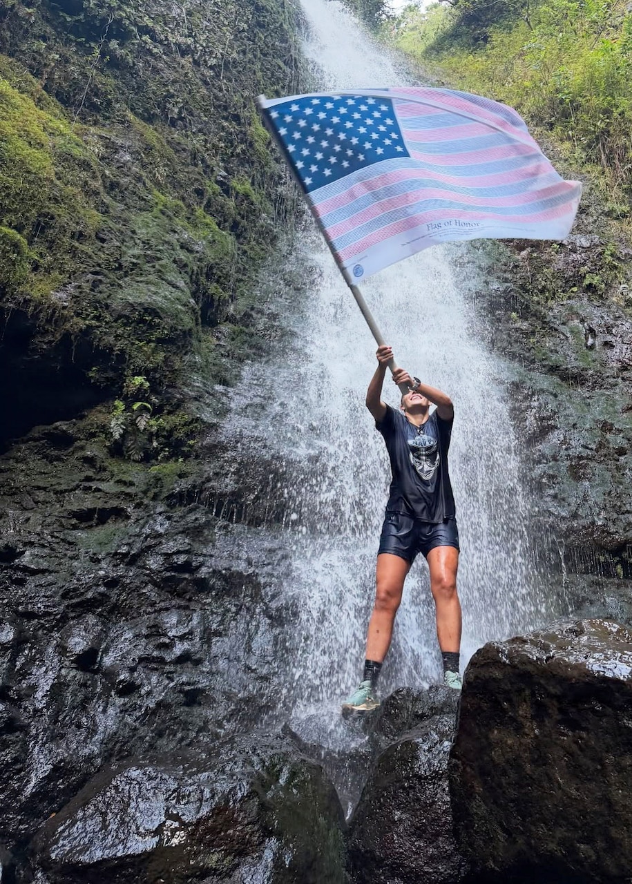 A woman holds a flag in the air and stands on a rock beneath a waterfall.