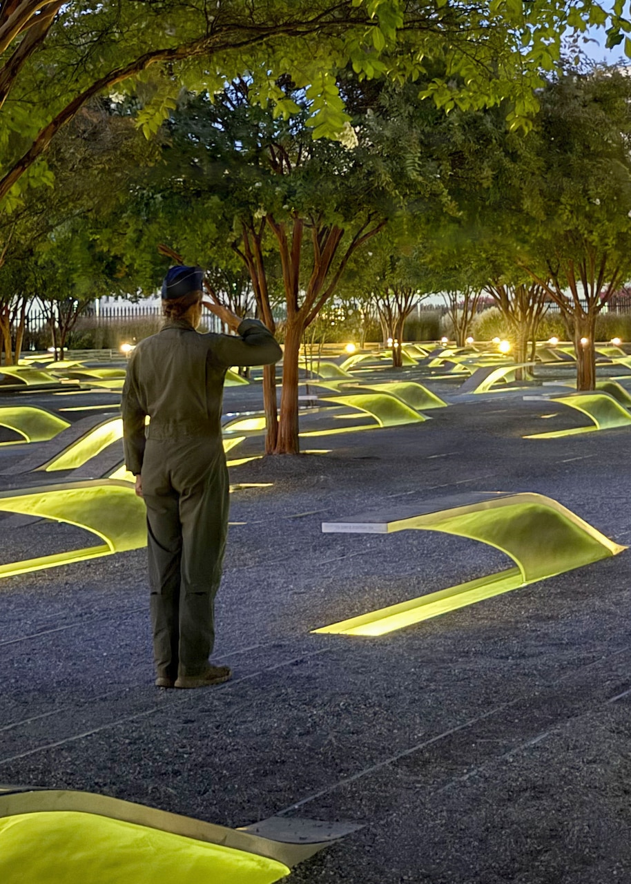 A woman in a flight suit stands and renders a salute in a memorial garden with trees and light-up benches at night.