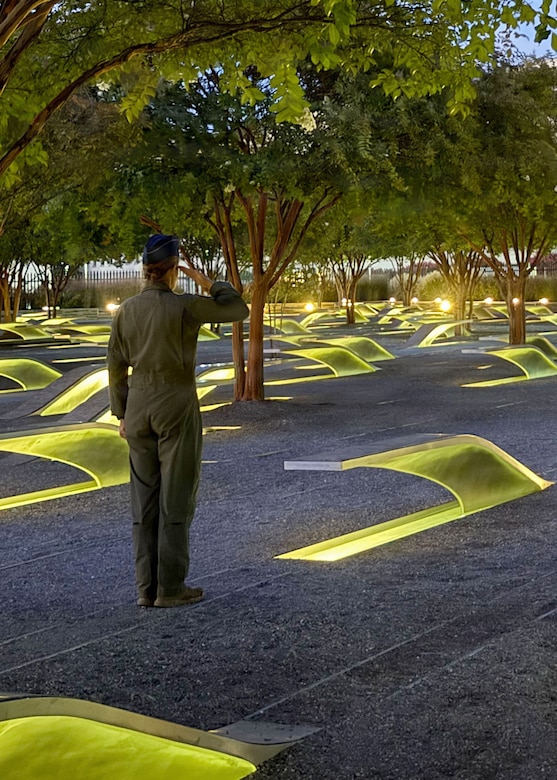 A woman in a flight suit stands and renders a salute in a memorial garden with trees and light-up benches at night.