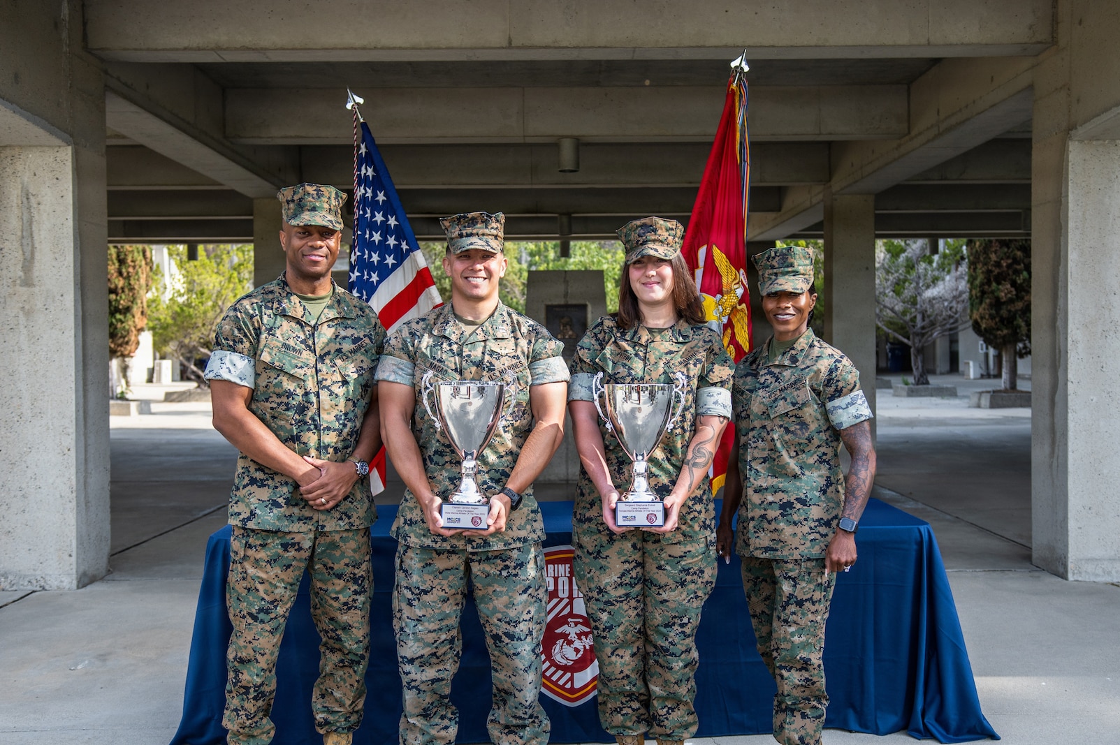 Four people in Marine Corps MARPAT uniforms pose for a photo with two tropies.