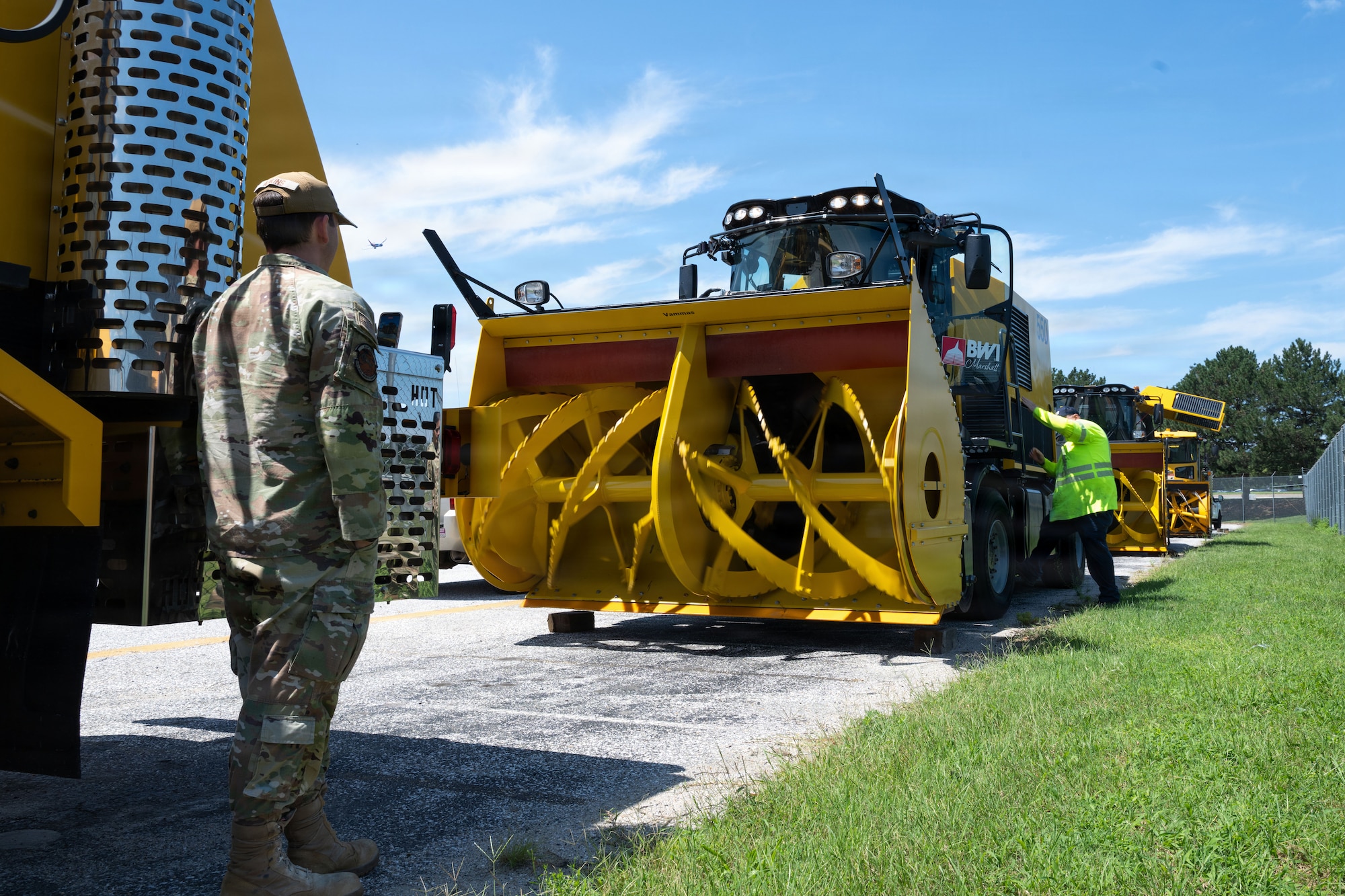 An airmen stands in front of a large yellow snow removal vehicle.