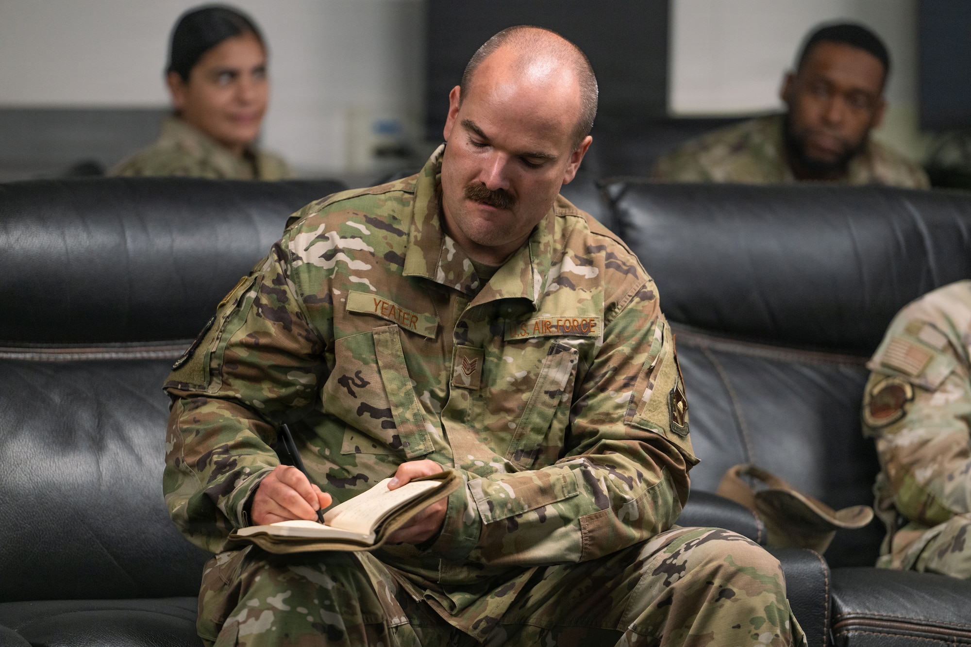 A man sits on a black sofa and writes in a a notebook.