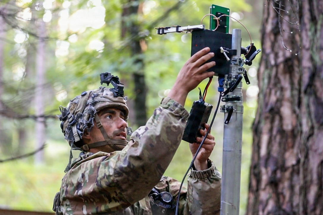 A soldier in tactical gear pushes buttons on a machine connected to a pole in a wooded area during the day.