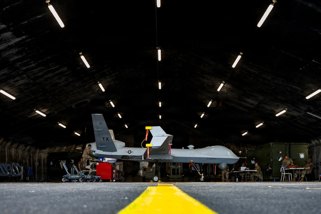A ground view of a yellow strip in the foreground and airmen working on an aircraft illuminated by hangar lights in the distance.