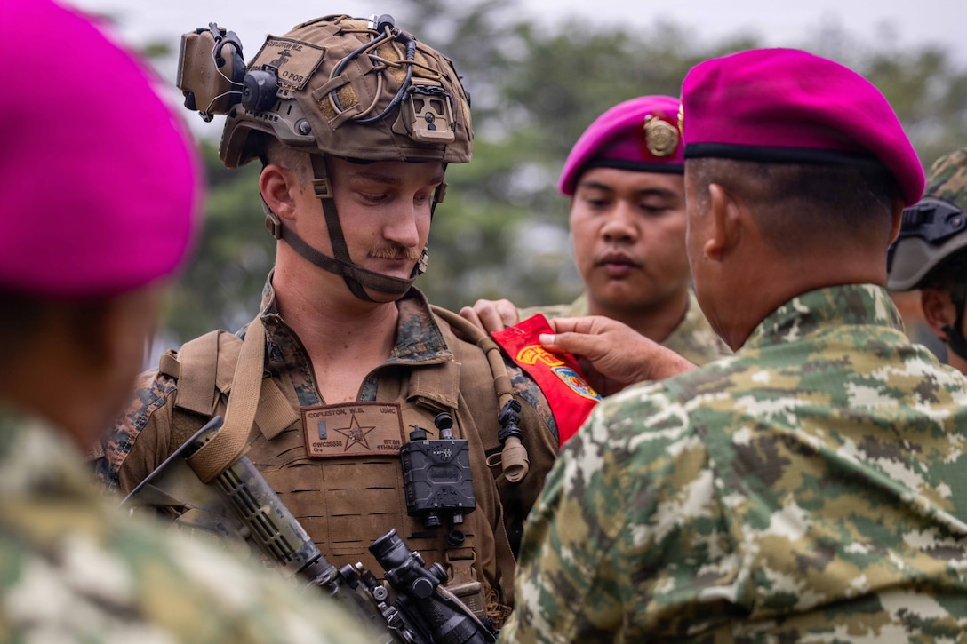 A U.S. Marine in tactical gear stands in the center as Indonesian marines in purple berets place a red patch on the U.S. service member's shoulder during the day.