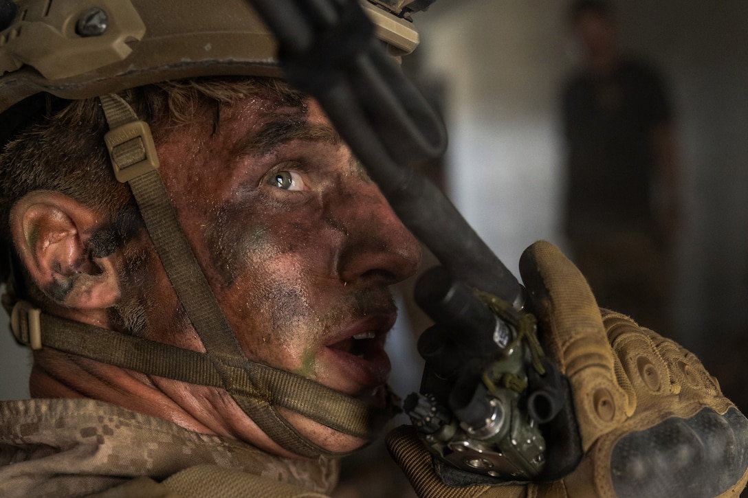 A Marine in a camouflage uniform and face paint looks to his right and speaks into a handheld device.