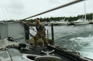A man in a camouflage military uniform speaks into a headset while standing on the side of a docked boat.