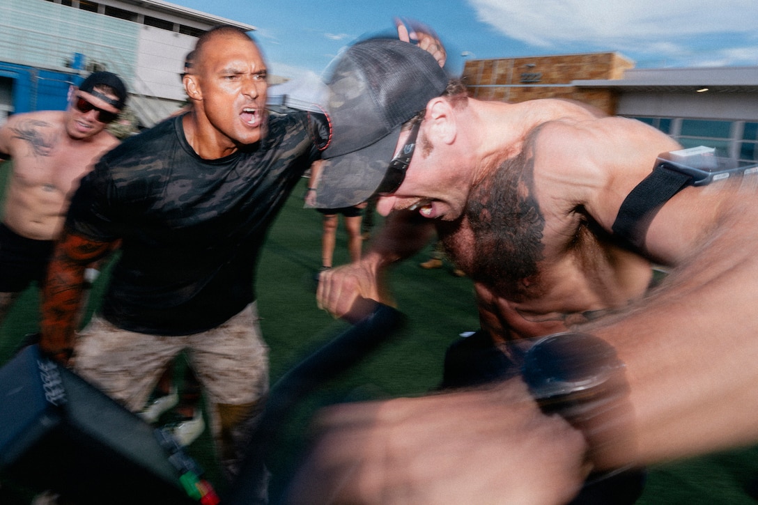 A sailor in athletic attire sans shirt rides an exercise bike on a field as another person in athletic attire shouts.
