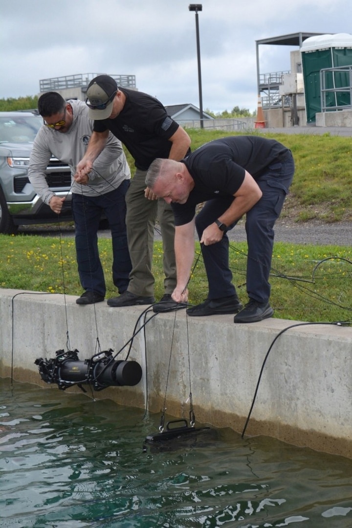 Photograph showing three men operating underwater x-ray near a concrete dock edge. The men are focused on lowering and maneuvering underwater x-ray connected by cables into a body of water, with grassy area and industrial structures in the background.