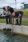 Photograph showing three men operating underwater x-ray near a concrete dock edge. The men are focused on lowering and maneuvering underwater x-ray connected by cables into a body of water, with grassy area and industrial structures in the background.