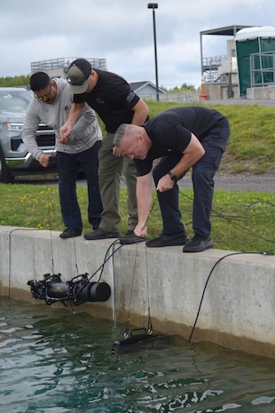 Photograph showing three men operating underwater x-ray near a concrete dock edge. The men are focused on lowering and maneuvering underwater x-ray connected by cables into a body of water, with grassy area and industrial structures in the background.