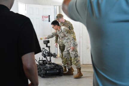 Photograph showing two military EOD Technicians in camouflage uniforms pointing/discussing oberservations over a tracked robot inside a building. The robot has a mounted camera and mechanical arm, with two other people partially visible from the back observing the discussion.