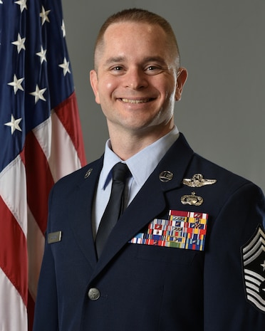 Chief Sjoholm poses for an official photo in his service dress. He is shown from the waist up with the U.S. flag over his right shoulder and a grey background.