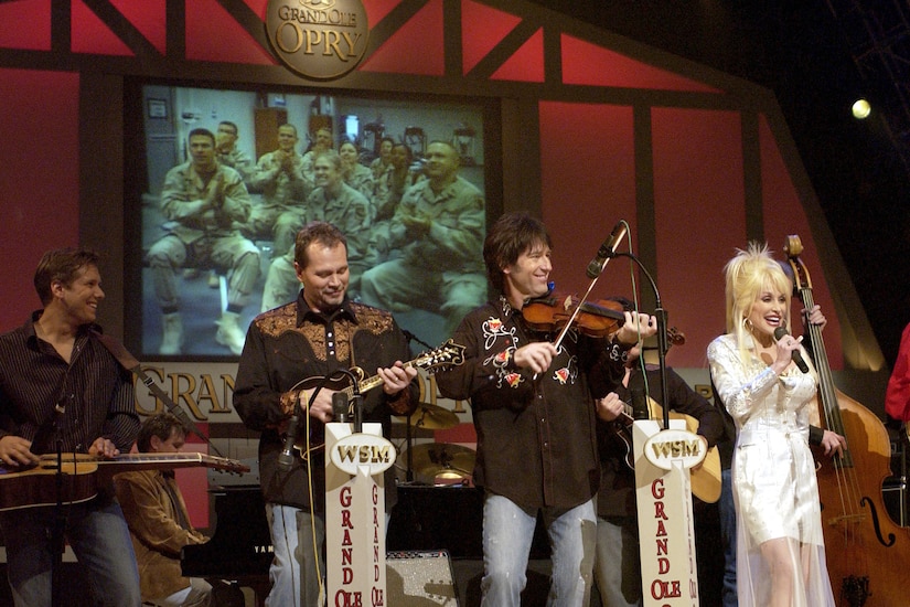 A woman in formal attire stands on a stage and sings into a microphone with a band behind her. Projected on a screen on the back wall are people in camouflage military uniforms sitting, smiling and clapping.