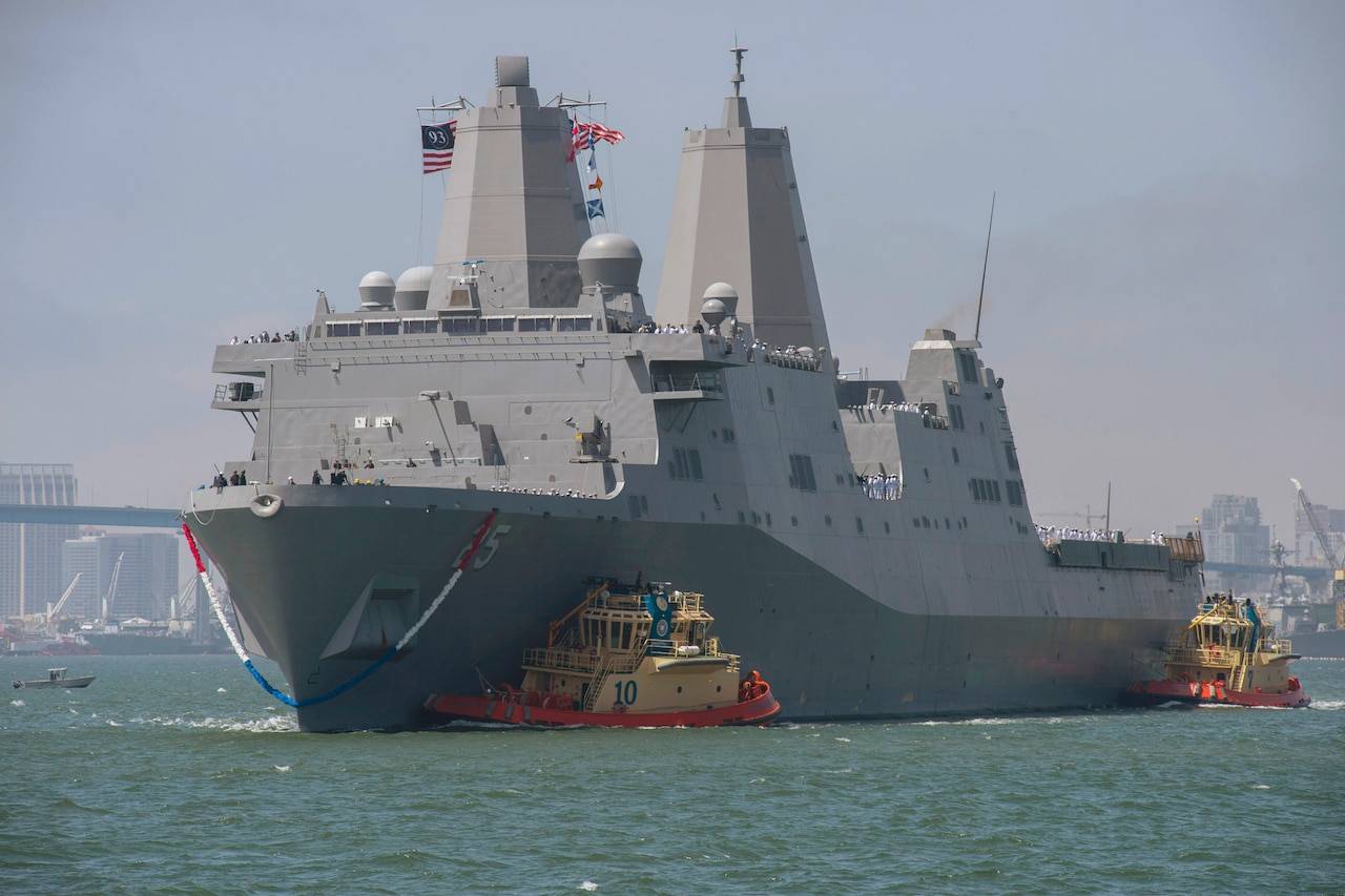 A ship with a 93  on an American flag comes into a port with two small yellow boats by its side.
