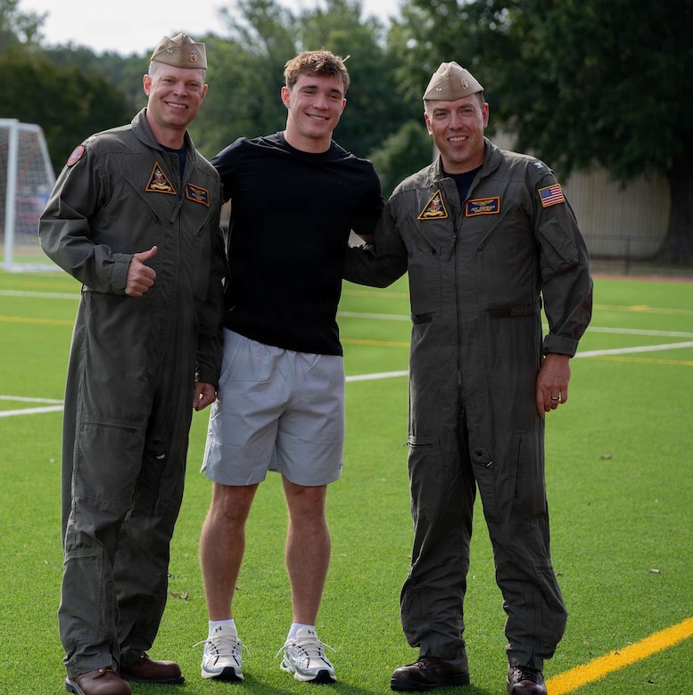 Two men in military flight suits smile as they stand next to a man in black and gray athletic attire.