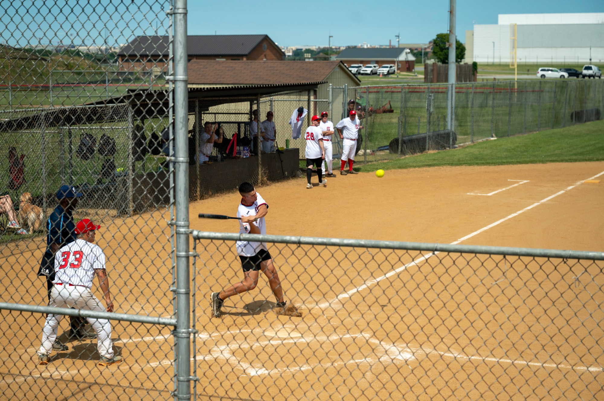 A softball player swings at a pitch during a game as teammates and players from both teams watch from the field and dugout.