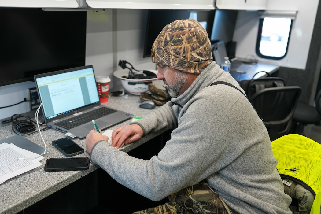 a man sits writing on a note pad while his laptop is open next to him.