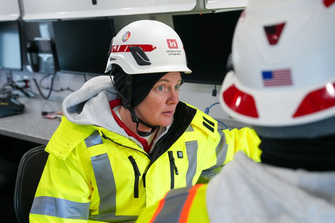 A woman in a hard hat and reflective coat sits looking at another individual who's back is to the camera.