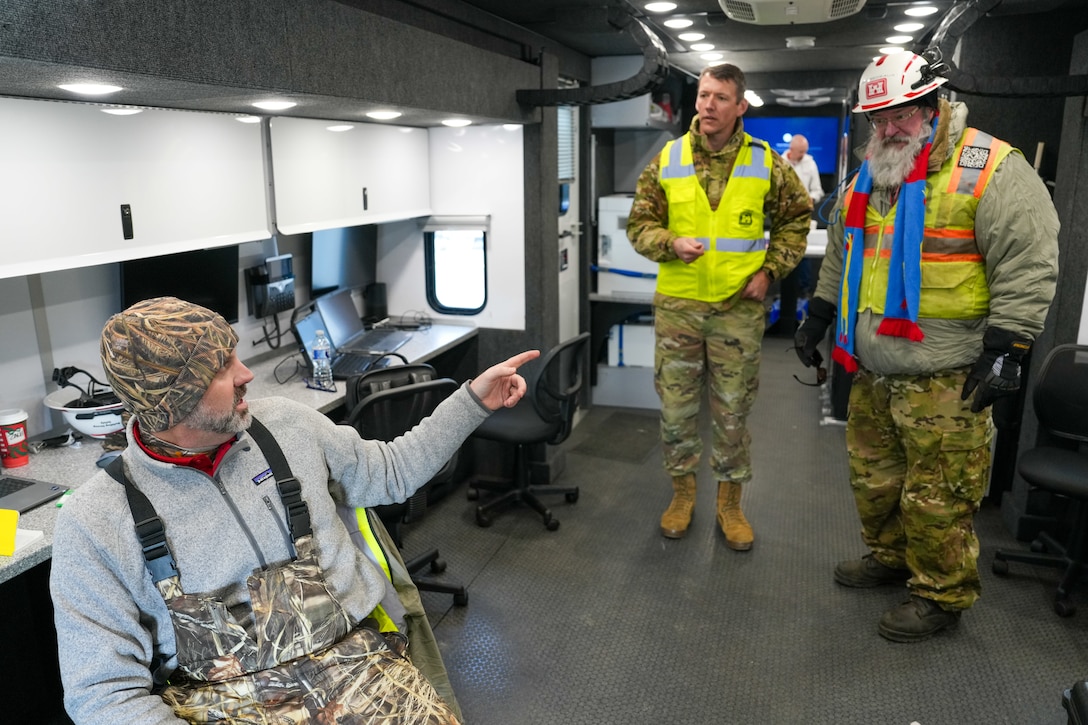 a man sitting in a chair talks with a soldier and another man who are standing near by.