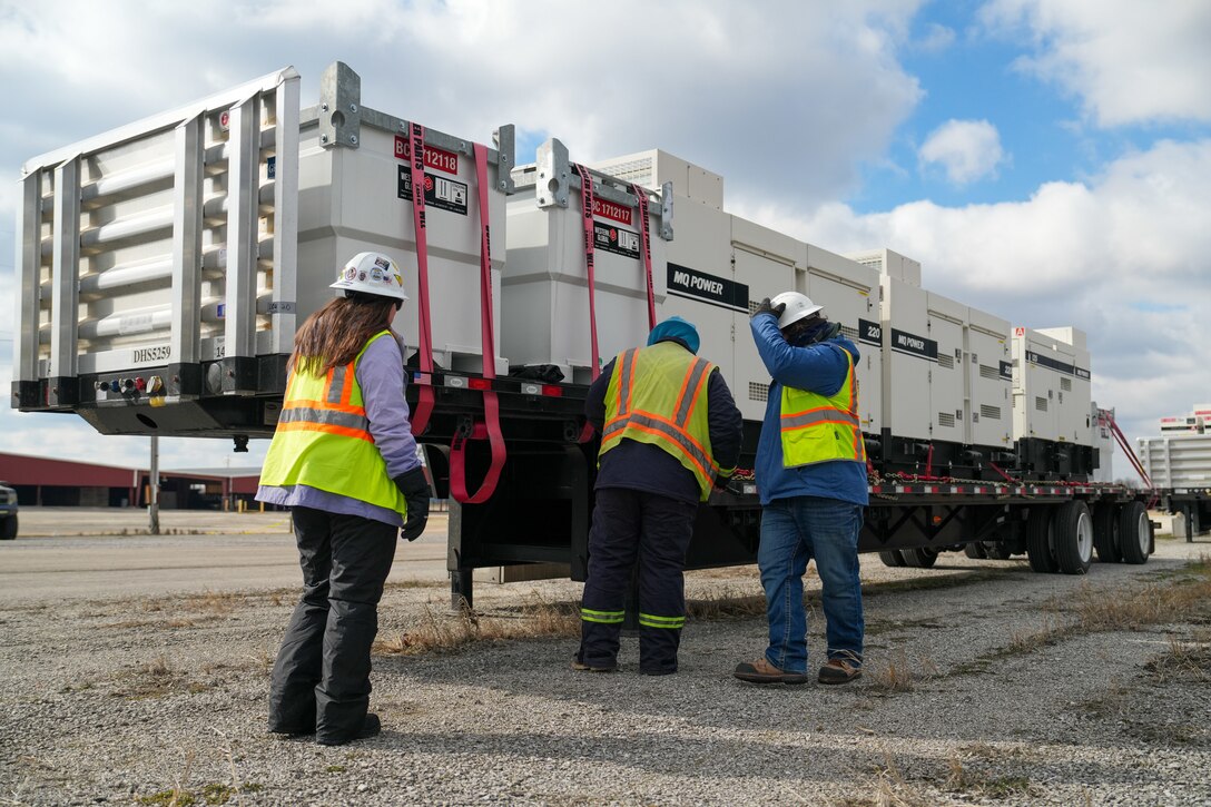 Three individuals inspect a large flatbed trailer loaded with generators and external fuel tanks.