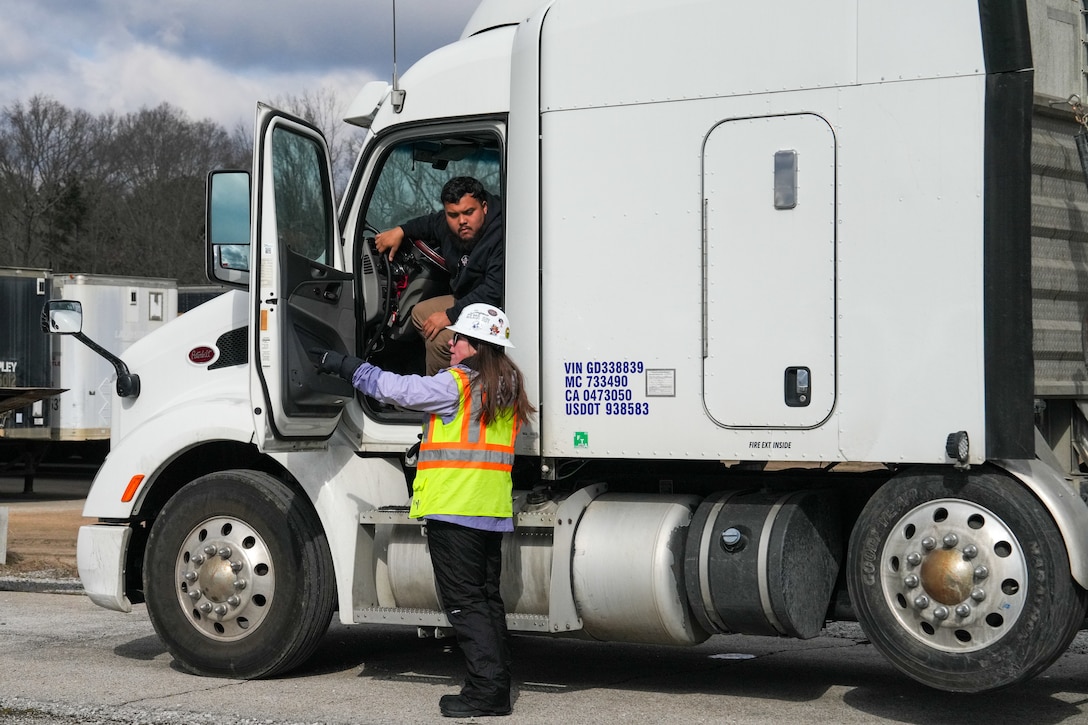 a woman in a hard hat and a safety vest talks to a tractor trailer driver who is sitting in his truck with the door open.