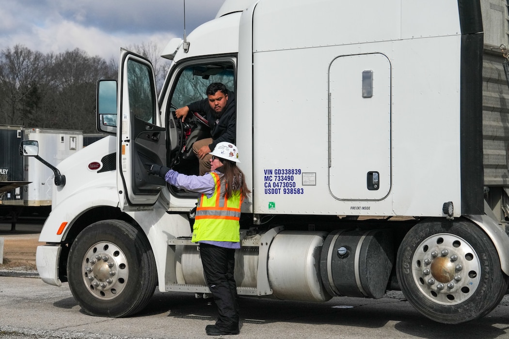 a woman in a hard hat and a safety vest talks to a tractor trailer driver who is sitting in his truck with the door open.