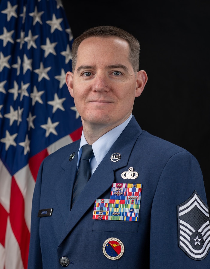 Official Photo of SMSgt Gregory Pflugh, United States Air Force Bands. SMSgt Pflugh is wearing navy blue service dress in front of an American flag.