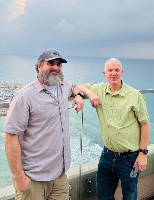 Two men posting by a fence overlooking water.