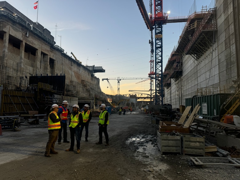 A group of men in protective gear talk to each other while construction is occurring around them.