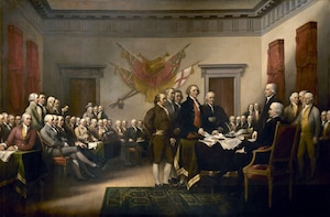 Men in historical attire stand around a table filled with documents, as others are seated or standing in a room with a high ceiling. 