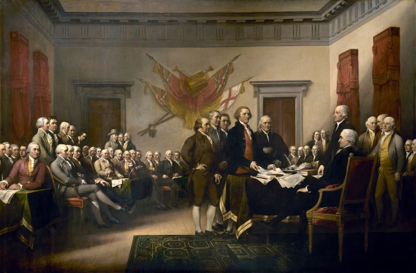 Men in historical attire stand around a table filled with documents as others in the distance are seated or standing in a room with a high ceiling.