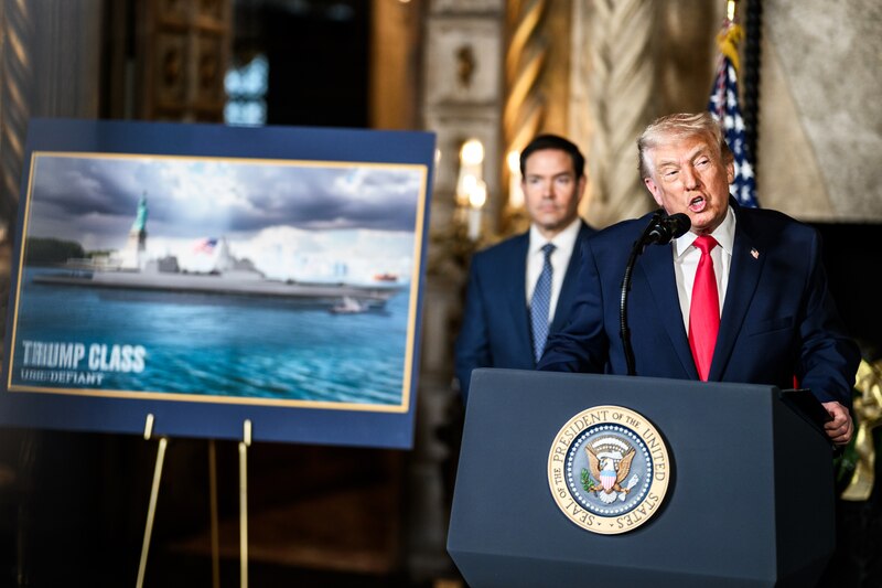 A man in a suit with a red tie talks from a lectern indoors as another man stands behind him.
