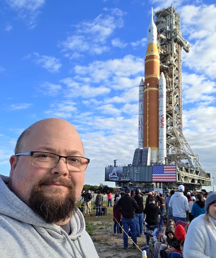 Man in the foreground poses for a selfie with a rocket ship on a platform in the background.