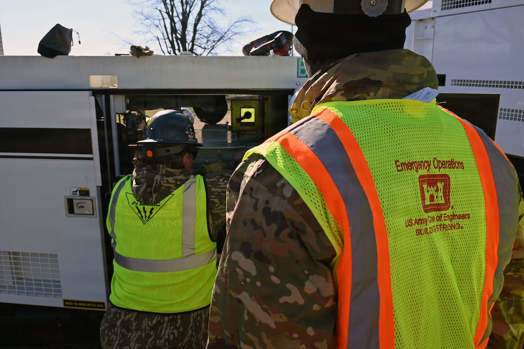 A soldier in a hard hat and reflective U.S. Army Corps of Engineer vest watches as a man works on a generator