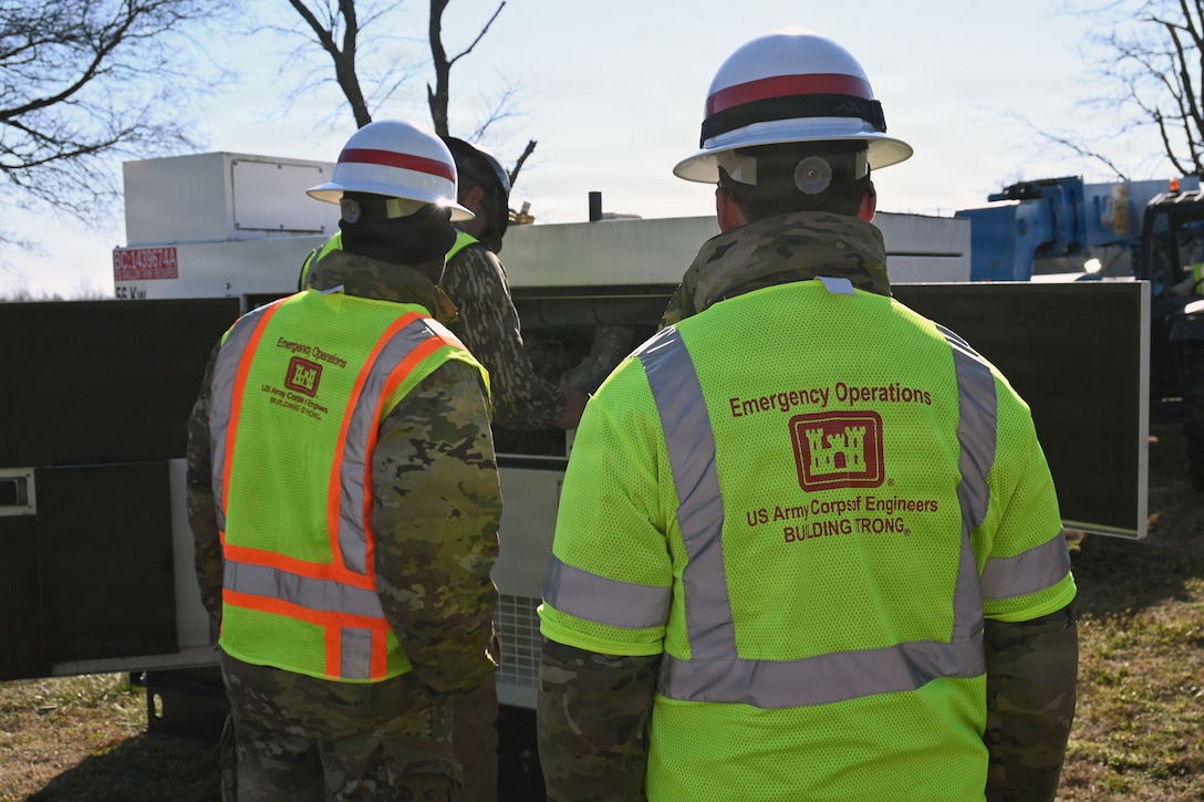 Two soldiers in hard hats and reflective U.S. Army Corps of Engineer vest watch as a man works on a generator