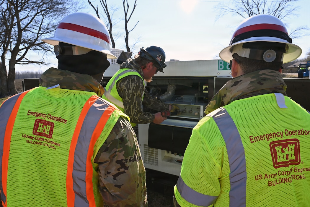 Two soldiers in hard hats and reflective U.S. Army Corps of Engineer vest watch as a man works on a generator