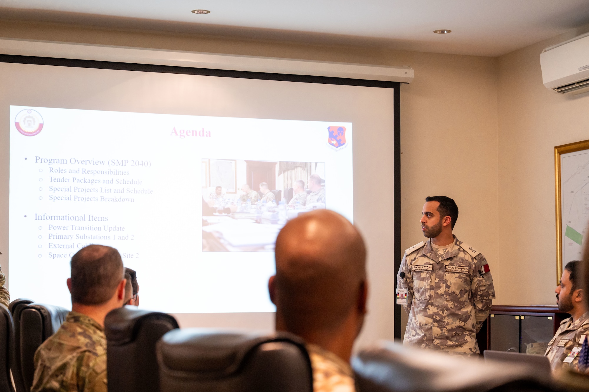 Lt Mohammed stands to the right of a projector screen and looks at it while people sit and listen to his brief