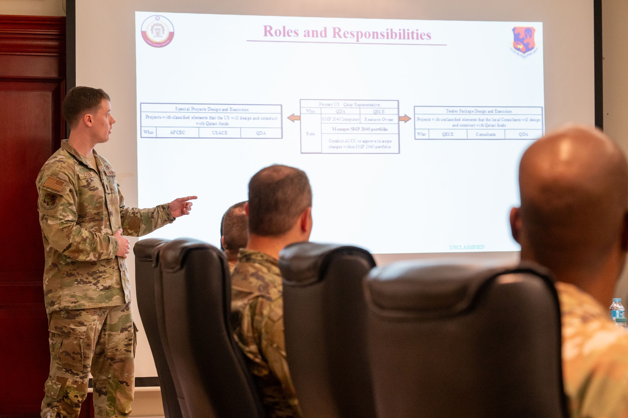 Lt Cross stands to left of a projector screen pointing at the presentation while people sit in chairs in the foreground