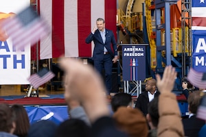 A person in civilian business attire salutes people from a stage setting; some in the crowd are waving small American flags.