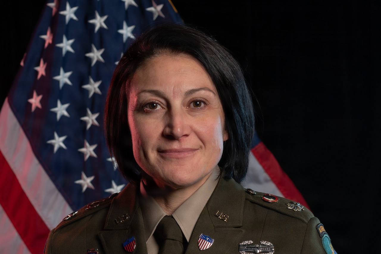 A woman wearing a military uniform poses for a photo in front of a flag.